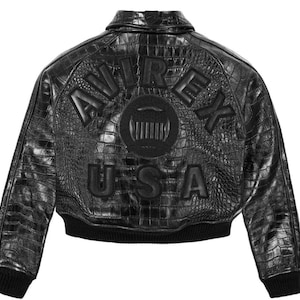 May include: A black leather jacket with a textured crocodile skin pattern. The back features the embossed text AVIREX USA in large letters and a circular emblem in the center. The jacket has a classic collar and black ribbed cuffs and hem.