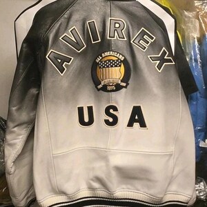 May include: A black and white leather jacket featuring a gradient fade design. The back displays the text AVIREX in large letters, a circular patch with the text ALL AMERICANS ESTABLISHED 1975 and a shield graphic, and the text USA below the patch.