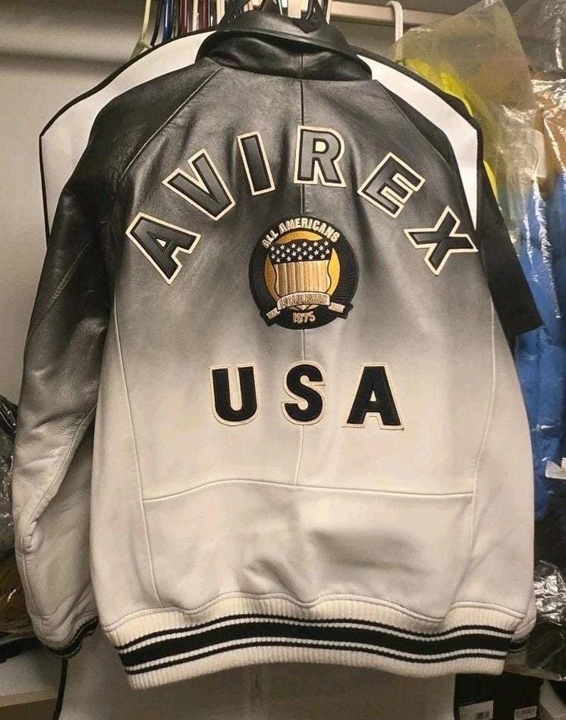 May include: A black and white leather jacket featuring a gradient fade design. The back displays the text AVIREX in large letters, a circular patch with the text ALL AMERICANS ESTABLISHED 1975 and a shield graphic, and the text USA below the patch.