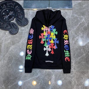 Könnte beinhalten: Schwarzer Kapuzenpullover mit farbenfrohem Kreuzdesign auf Rücken und Ärmeln. Die Ärmel zeigen das Wort "CHROME HEARTS" in einem Regenbogenverlauf. Der Hoodie hat eine Kapuze mit Kordelzug und eine Kängurutasche.