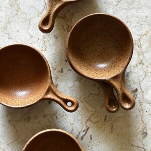May include: A set of four round ceramic bowls with handles, each featuring a speckled brown glaze. The bowls are arranged in a cluster, showcasing their circular shape, deep interior, and small handles with a hole for hanging.