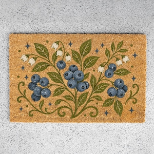 May include: A rectangular doormat with a botanical design featuring blueberries, green leaves, and white flowers. The design is set against a natural coir background, with small blue star accents.