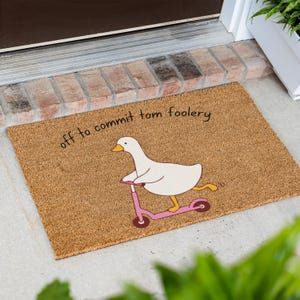 May include: A rectangular doormat made of natural fibers. It features a cartoon goose riding a pink scooter, with the phrase "off to commit tom foolery" in black script. The mat is placed in front of a door.