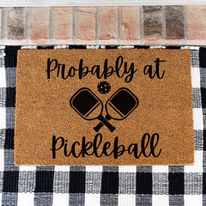 May include: A rectangular tan coir doormat featuring the black text "Probably at Pickleball" and a graphic of two crossed pickleball paddles with a ball in the center. The mat has a coarse, natural fiber texture and a clean, simple design.