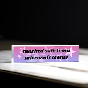 May include: A rectangular acrylic desk sign featuring a pink and purple gradient background with white star outlines and the black text "marked safe from microsoft teams" printed across the center of the front face of the clear block.