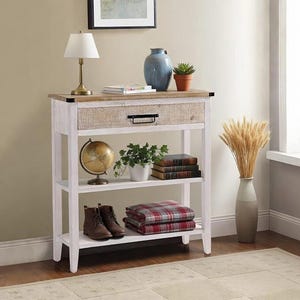 May include: A white and wood-tone console table with a drawer and two shelves. The top shelf holds a lamp, vase, and plant. The middle shelf has a globe, plant, and books. The bottom shelf displays boots and a plaid blanket.