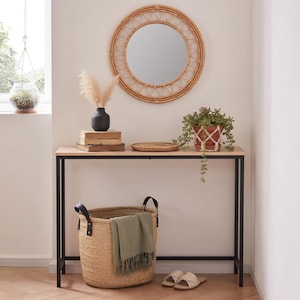 May include: A round rattan mirror hangs above a console table with a black metal frame and a light wood top. A woven basket with a green blanket sits below. The table is decorated with a black vase, books, a small plant, and a woven tray. Beige sandals are on the floor.