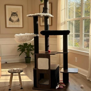 May include: A multi-level cat tree with a dark brown and black color scheme. It features multiple platforms, a plush bed, scratching posts, and a hanging toy. The cat tree is placed in a room with a framed cat portrait and a small stool.
