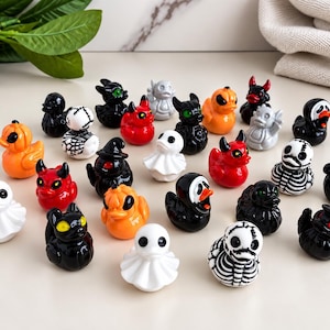 May include: A collection of small, decorative rubber ducks featuring various Halloween-themed designs, including ghosts, skeletons, pumpkins, devils, and black cats. Each miniature figure is painted in vibrant colors like orange, red, black, and white.