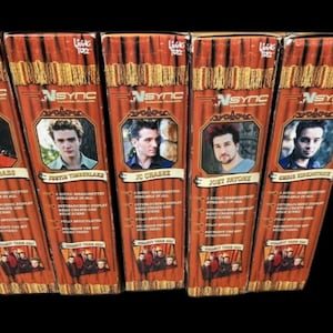 Op de afbeelding: Vijf verzameldozen met *NSYNC bandleden: Lance Bass, Justin Timberlake, JC Chasez, Joey Fatone en Chris Kirkpatrick. Elke doos heeft een portret van een bandlid en het *NSYNC logo. De dozen zijn bruin met een podiumgordijn ontwerp.