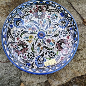 May include: A round, hand-painted ceramic plate with a vibrant floral design. The plate features a white background with blue, pink, yellow, and green floral patterns. The rim is a deep blue with a decorative border.