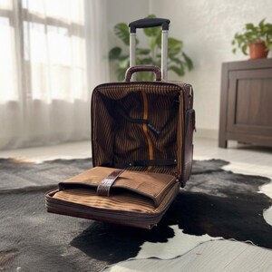 Op de afbeelding: Bruine rolkoffer met een open compartiment dat een gestreept interieur onthult. De koffer heeft een telescopische handgreep en een handvat aan de bovenkant. De buitenkant lijkt van getextureerd leer te zijn. De koffer staat op een zwart-wit koeienvachtkleed.