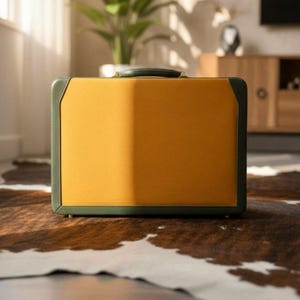 May include: A rectangular suitcase with a mustard yellow front and a green trim. The suitcase has a green handle and is sitting on a brown and white cowhide rug. The suitcase is a neutral color and has a classic design.