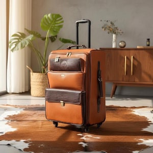 Könnte beinhalten: Ein brauner Rollkoffer mit mehreren Außentaschen. Der Koffer hat einen Teleskopgriff und besteht aus einem strukturierten Material. Die Taschen sind dunkler braun mit einer Krokodilleder-Textur. Der Koffer steht auf einem braun-weißen Teppich.