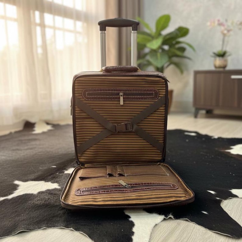 Op de afbeelding: Bruin en tan rollende aktetas met een telescopische handgreep. De aktetas heeft een gestreept patroon en bruine lederen accenten. Het interieur is open en toont compartimenten en een vak met ritssluiting. De koffer staat op een zwart-wit koeienvacht tapijt.