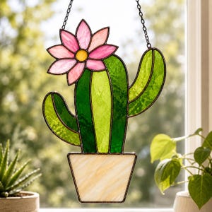 以下が含まれることがあります： 鉢植えのサボテンの形をしたステンドグラスのサンキャッチャー。サボテンは様々な色合いの緑色のガラスで作られており、上部には黄色い中心を持つピンクの花があしらわれています。鉢はクリーム色のガラス製で、金属製のチェーンで吊り下げられています。
