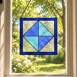 May include: A square stained glass suncatcher featuring a geometric pattern of four triangles in alternating shades of deep blue and light turquoise. The piece is framed in dark blue glass and suspended by a black metal chain for hanging.