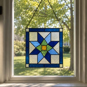 May include: A square stained glass panel featuring a central yellow square surrounded by green, light blue, and dark blue geometric star shapes. The panel is framed by dark blue borders and clear textured glass, suspended by a black metal chain.