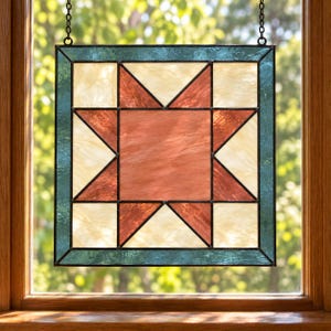May include: A square stained glass panel featuring a central eight-pointed star design in shades of terracotta and cream. The panel is framed by a border of textured teal glass and held by a black metal frame with hanging chains.