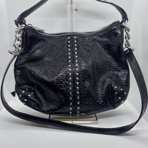 May include: Black patent leather shoulder bag with silver-tone hardware. The bag features a zip closure, studded accents, and a chain detail on the straps. The bag has a long strap for shoulder or crossbody wear.