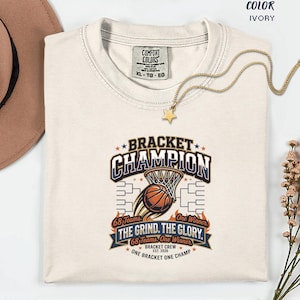May include: Ivory-colored t-shirt with a basketball-themed graphic. The design includes the words "BRACKET CHAMPION," a basketball going into a net, and the text "THE GRIND. THE GLORY." The shirt is a Comfort Colors XL-TG-EG.