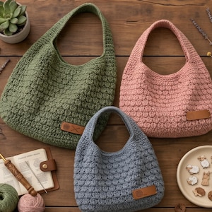 May include: Three handmade crochet bags in olive green, dusty rose, and slate blue, each with a textured stitch pattern and a leather tag. The bags are displayed on a wooden surface, alongside crochet supplies and decorative elements.