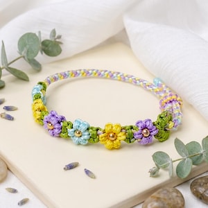 May include: A colorful bracelet featuring a braided band in pastel hues of yellow, purple, and green. The bracelet is adorned with small, embroidered flower accents in yellow, purple, and blue, each with a gold bead center. The bracelet is a delicate piece of jewelry.