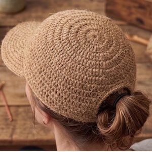 May include: A tan crocheted baseball cap with a curved brim. The cap is made of a textured, woven material and has a circular pattern on top. The cap is worn with the hair pulled back in a low bun.