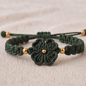 May include: Dark green crochet bracelet with a four-leaf clover design. The bracelet features gold-coloured beads and an adjustable sliding knot closure. The clover is the focal point, with detailed stitching and a central gold bead.