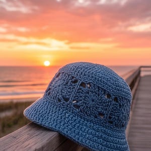 Peut inclure: Un chapeau bleu crocheté avec un motif floral est posé sur une rambarde en bois. Le chapeau a un bord et des découpes décoratives. L'arrière-plan présente un coucher de soleil sur la mer, avec une passerelle en bois.