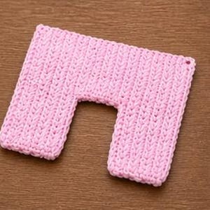 May include: A light pink, U-shaped, knitted neck wrap. The wrap is rectangular with a curved cutout in the center. The knitted texture is visible, and the wrap is lying on a wooden surface.