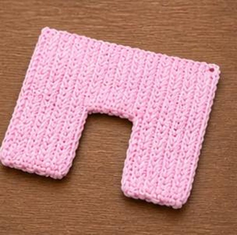 May include: A light pink, U-shaped, knitted neck wrap. The wrap is rectangular with a curved cutout in the center. The knitted texture is visible, and the wrap is lying on a wooden surface.