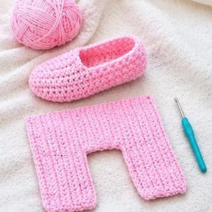May include: A pink crocheted slipper, a rectangular crocheted piece, a ball of pink yarn, and a blue crochet hook are arranged on a white surface. The slipper and rectangular piece are made of the same pink yarn. The items suggest a crafting project.