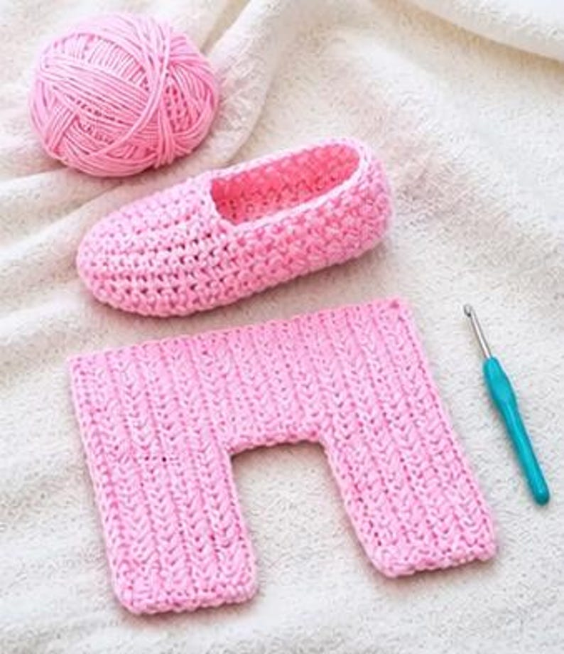 May include: A pink crocheted slipper, a rectangular crocheted piece, a ball of pink yarn, and a blue crochet hook are arranged on a white surface. The slipper and rectangular piece are made of the same pink yarn. The items suggest a crafting project.
