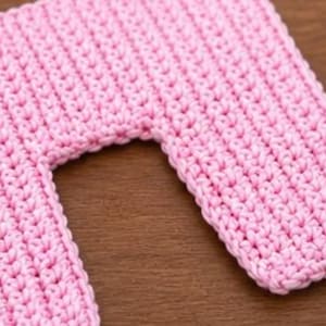 May include: Close-up of a pink crocheted item with a rectangular shape and a curved cutout. The crochet stitches are visible, creating a textured surface. The item is likely handmade and sits on a wooden surface.