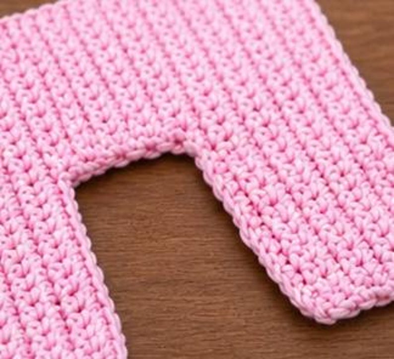 May include: Close-up of a pink crocheted item with a rectangular shape and a curved cutout. The crochet stitches are visible, creating a textured surface. The item is likely handmade and sits on a wooden surface.