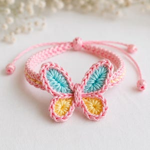 May include: A pink crochet bracelet featuring a butterfly design. The butterfly has blue and yellow accents, with a pink border. The bracelet is adjustable with a sliding knot closure, and is a delicate accessory.