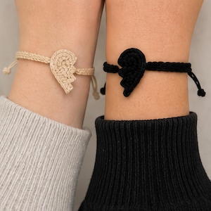 May include: Two matching crochet bracelets, each featuring a half-heart design. One bracelet is beige, the other is black. The bracelets are worn on wrists, with adjustable ties for a comfortable fit. The bracelets are a symbol of friendship.
