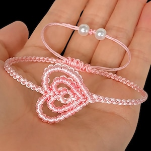 May include: Two pink bracelets. One features a heart design, the other has two white pearls. Both are made of woven pink cord. The bracelets are displayed against a neutral background.
