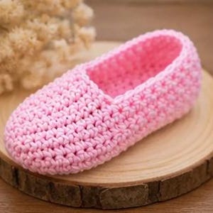 May include: A handmade pink crochet slipper. The slipper is made of soft yarn and has a textured stitch pattern. It is displayed on a round wooden surface, with a blurred background of dried flowers.