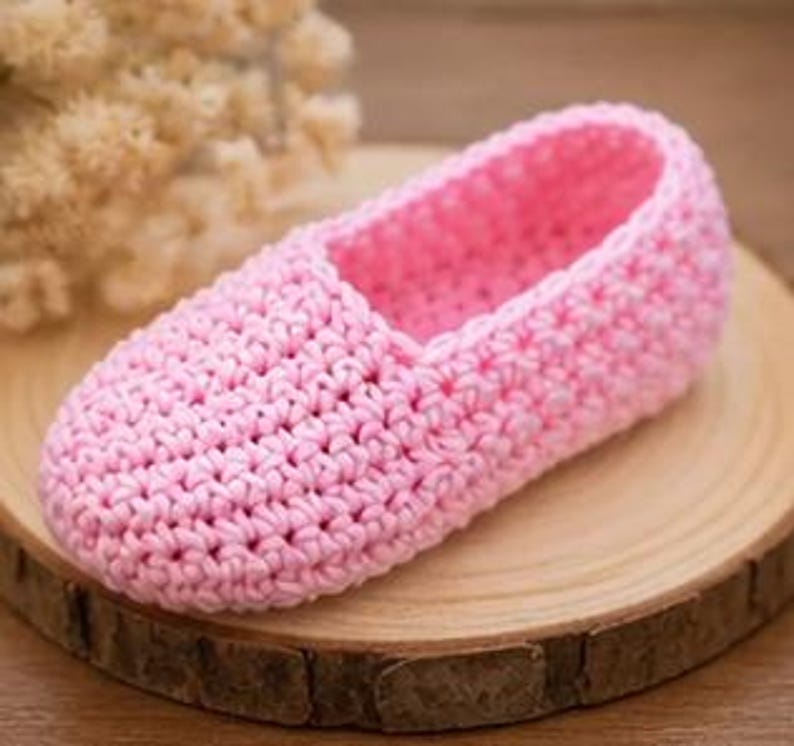 May include: A handmade pink crochet slipper. The slipper is made of soft yarn and has a textured stitch pattern. It is displayed on a round wooden surface, with a blurred background of dried flowers.