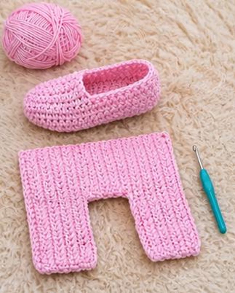 May include: A pink crochet slipper, a pink yarn ball, and a pink crochet rectangle are displayed on a textured surface. A teal crochet hook is also present. The slipper is in the shape of a shoe. The rectangle has a U-shaped cutout.