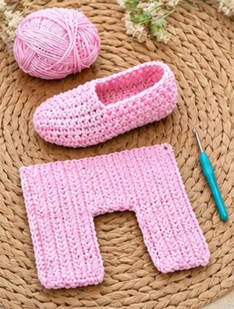 May include: A pink crocheted slipper, a matching rectangular piece, and a ball of pink yarn are arranged on a woven mat. A blue and silver crochet hook is also present. The slipper has a soft, fuzzy interior.