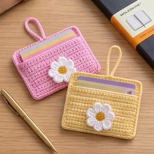 Peut inclure: Deux porte-cartes crochetés à la main, un rose et un jaune, chacun orné d'une marguerite blanche. Les porte-cartes ont une boucle pour faciliter le transport et sont remplis de cartes colorées. Un stylo doré repose sur la surface en bois.
