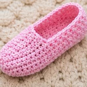 May include: A single, light pink crochet slipper. The slipper is made with a tight stitch pattern and has a rounded toe and a low-cut opening. The slipper is sitting on a cream-colored, textured surface.