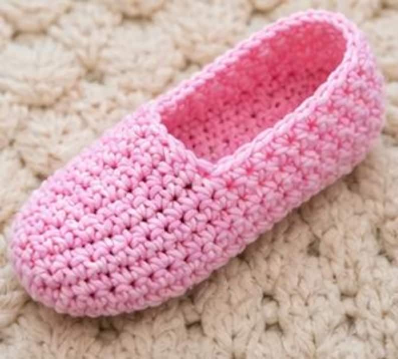 May include: A single, light pink crochet slipper. The slipper is made with a tight stitch pattern and has a rounded toe and a low-cut opening. The slipper is sitting on a cream-colored, textured surface.