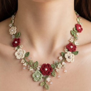 May include: A handcrafted floral necklace featuring crocheted flowers in cream, red, and green. The necklace has a delicate design with pearl accents and green leaves, creating a natural, elegant look. The necklace is a unique piece of jewelry.
