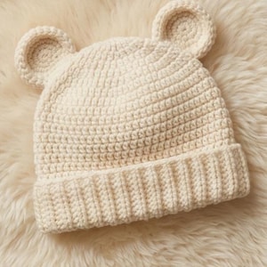 May include: A cream-coloured crochet hat with bear ears. The hat has a ribbed brim and a textured stitch pattern across the body and ears. It is displayed on a fluffy, cream-coloured surface.