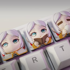 Może przedstawiać: Zbliżenie trzech niestandardowych nakładek na klawisze z postaciami w stylu anime z białymi włosami i niebieskimi oczami. Jedna nakładka przedstawia postać czytającą książkę, druga pije z kubka. Nakładki znajdują się na białej klawiaturze.