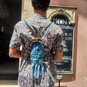 May include: A squid-shaped bag featuring a dark blue glittery head with teal accents and dangling light blue tentacles. The bag has a metal kiss-lock clasp at the top and is worn with thin teal braided straps over a patterned short-sleeve shirt.